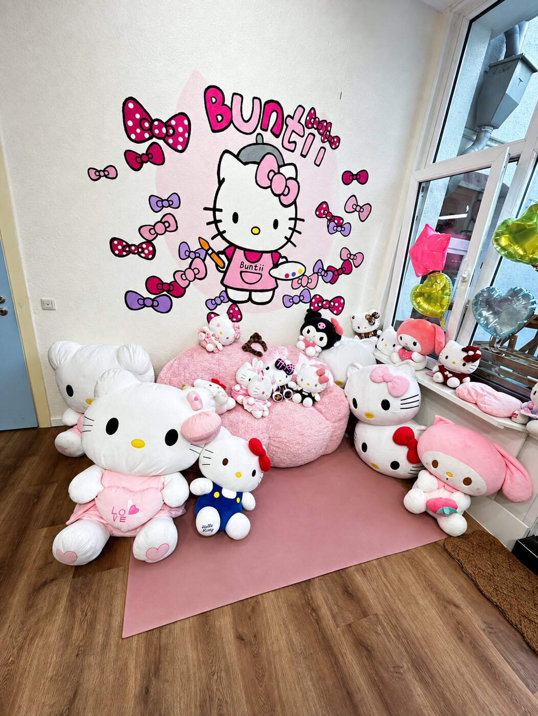 Fotoecke mit handgemaltem Hello-Kitty-Wandbild mit Pinsel und Palette, darunter riesige Plüschtiere auf rosa Teppich.