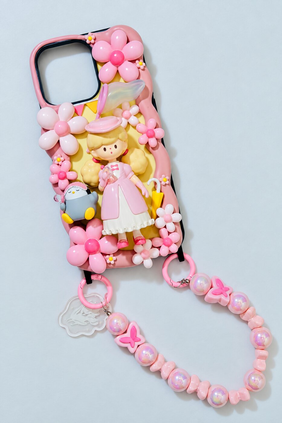 Fertige Deko-Handyhülle mit rosa Cremeblüten, Puppenfigur, Pinguin-Anhänger und rosa Perlenkette.